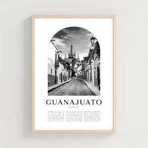 Op de afbeelding: Zwart-wit ingelijste print van Guanajuato, Mexico. De afbeelding toont een geplaveide straat die naar een kerk leidt, omlijst door een boog. Het woord "GUANAJUATO" is vetgedrukt onder de afbeelding afgedrukt.