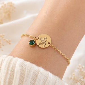 Könnte beinhalten: Ein goldfarbenes Kettenarmband mit einem runden Anhänger, der mit einem Blumenmotiv und dem Buchstaben J graviert ist, sowie einem kleinen, runden, dunkelgrünen facettierten Steinanhänger. Das Armband besteht aus einer feinen Gliederkette.
