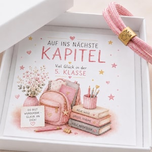 Könnte beinhalten: Eine weiße Geschenkbox mit einem rosa Kordelarmband, das eine goldfarbene Metallperle mit der Gravur Mia und einem Stern enthält, sowie einer Karte mit Illustrationen von Rucksack, Büchern und Blumen mit dem Text Auf ins nächste Kapitel.