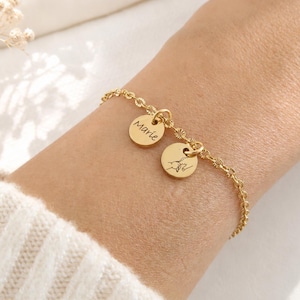 Könnte beinhalten: Ein goldfarbenes Kettenarmband mit zwei kleinen runden Anhängern. Ein Anhänger ist mit dem Namen Marie in Schreibschrift graviert, während der andere eine Strichzeichnung von zwei Händen zeigt, die sich an den Fingern halten.