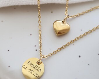 May include: Two gold-colored necklaces. One features a heart pendant, the other a round pendant engraved with the name "Emilia" and the date "01.08.22". The necklaces are displayed on a white surface.