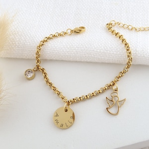 May include: Gold bracelet featuring a clear crystal charm, a disc pendant engraved with "Amalia", and an angel silhouette. The bracelet is displayed on a white background.
