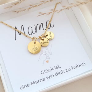 May include: A gold-toned necklace featuring three small circular charms attached to a delicate chain. One charm displays an engraved line drawing of two hands holding fingers, while the other two charms are engraved with the names Mia and Leon.