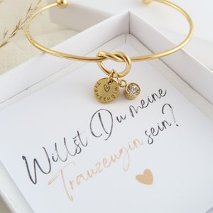Könnte beinhalten: Ein goldfarbenes Armband mit einem Knoten-Design, einem kleinen runden Anhänger mit Herz-Gravur und dem Text "Trauzeugin" sowie einem klaren Kristallanhänger, präsentiert in einer weißen Schachtel mit dem Text "Willst Du meine Trauzeugin sein?"