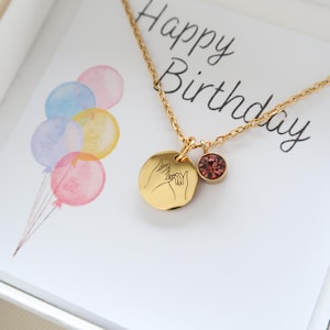 May include: Gold necklace with a pink crystal charm and a round pendant with an engraved image of two hands making a pinky promise. The necklace is in a white box with a card that says "Happy Birthday".