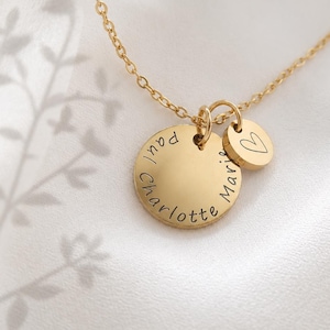 May include: A gold-toned necklace featuring a circular pendant engraved with the names Paul, Charlotte, and Marie, accompanied by a smaller circular charm displaying a simple heart outline, all suspended from a delicate gold-toned chain.