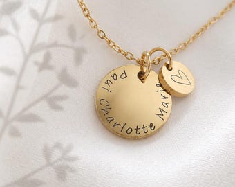 Könnte beinhalten: Eine goldfarbene Halskette mit einem runden Anhänger, in den die Namen Paul, Charlotte und Marie eingraviert sind, ergänzt durch einen kleineren runden Anhänger mit einem Herzumriss, befestigt an einer feinen goldfarbenen Kette.