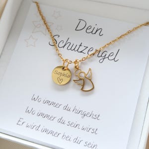 May include: A gold-colored necklace with two charms, one a round disc engraved with "Sophia" and a heart, the other an angel silhouette. The necklace rests on a card with German text, including "Dein Schutzengel."