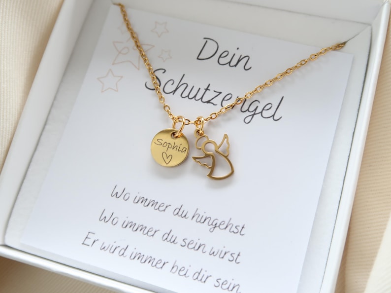 May include: A gold-colored necklace with two charms, one a round disc engraved with "Sophia" and a heart, the other an angel silhouette. The necklace rests on a card with German text, including "Dein Schutzengel."