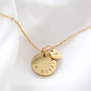 May include: A gold-colored necklace with two circular pendants. The larger pendant has the names "Mia" and "Sophie" engraved on it. The smaller pendant features a hand gesture. The necklace is on a white background.