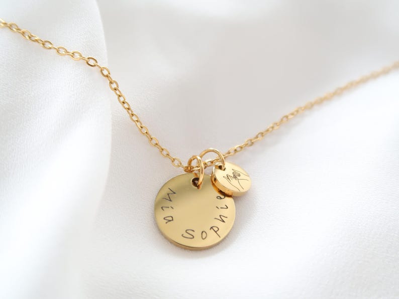 May include: A gold-colored necklace with two circular pendants. The larger pendant has the names "Mia" and "Sophie" engraved on it. The smaller pendant features a hand gesture. The necklace is on a white background.