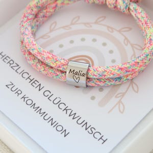 Könnte beinhalten: Ein mehrfarbiges geflochtenes Kordelarmband mit einer silberfarbenen Metallperle, die mit dem Namen Malia und einem Herzsymbol graviert ist. Das Armband liegt auf einer weißen Karte mit dem Text HERZLICHEN GLÜCKWUNSCH ZUR KOMMUNION.