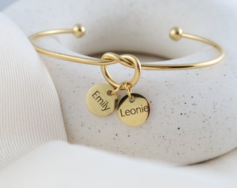 May include: Gold bangle bracelet with a knot charm and two round charms with the names "Emily" and "Leonie" engraved on them.