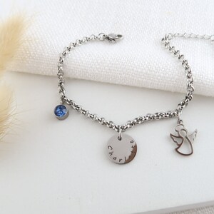 May include: A silver bracelet with a chain of round links. It has a blue gemstone, a disc charm engraved with "Charlotte", and an angel charm. The bracelet is shown on a white surface.