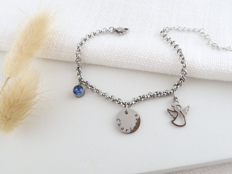 May include: A silver bracelet with a chain of round links. It has a blue gemstone, a disc charm engraved with "Charlotte", and an angel charm. The bracelet is shown on a white surface.