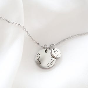 May include: A silver necklace with two circular pendants. The larger pendant has the name "Clara" and "Sophie" engraved on it. A smaller pendant with a heart symbol is also attached. The necklace is displayed on a white background.