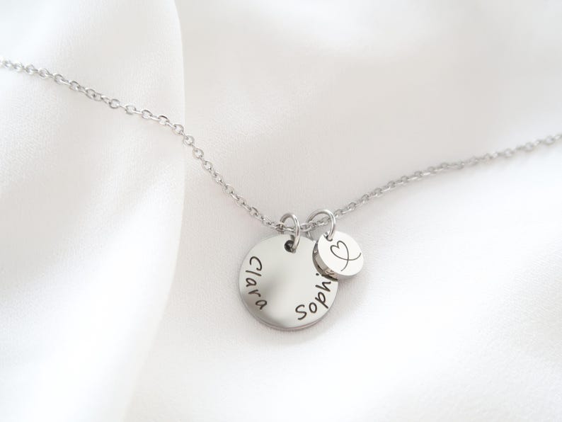 May include: A silver necklace with two circular pendants. The larger pendant has the name "Clara" and "Sophie" engraved on it. A smaller pendant with a heart symbol is also attached. The necklace is displayed on a white background.