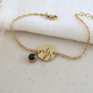 May include: Gold-toned bracelet featuring a round charm with a floral design and the letter "J". A small, round, emerald-green gemstone hangs from the chain. The bracelet is on a white surface.