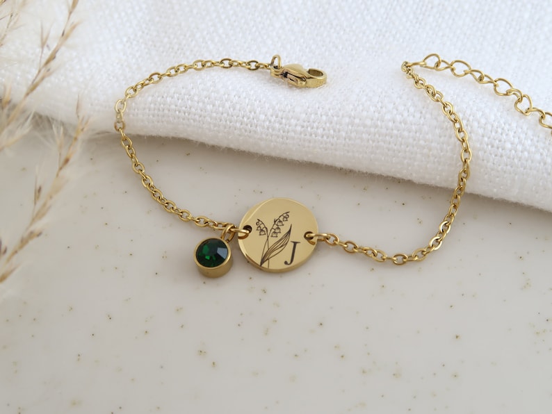 May include: Gold-toned bracelet featuring a round charm with a floral design and the letter "J". A small, round, emerald-green gemstone hangs from the chain. The bracelet is on a white surface.