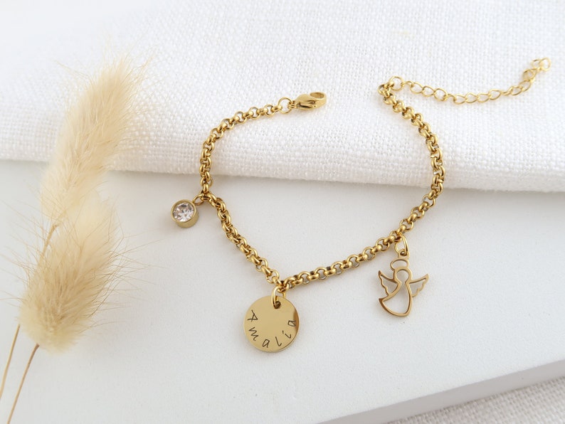 May include: Gold bracelet featuring a clear crystal charm, a disc pendant engraved with "Amalia", and an angel silhouette. The bracelet is displayed on a white background.