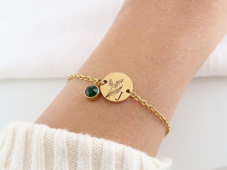 May include: Gold-toned bracelet featuring a round charm with a floral design and the letter "J". A small, round green gemstone hangs next to the charm. The bracelet is on a chain.