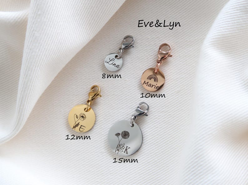 Op de afbeelding: Vier ronde metalen bedels met karabijnsluitingen in zilver-, goud- en ros&eacute;goudkleuren. De bedels zijn gegraveerd met namen, letters en bloem- of regenboogontwerpen. Labels geven maten van 8mm, 10mm, 12mm en 15mm aan. De tekst Eve&Lyn is zichtbaar.