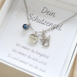 May include: A silver necklace with three charms, presented in a white box. The charms include a blue gemstone, a round pendant with the name "Emilia" and a heart, and a small angel. The card reads "Dein Schutzengel".