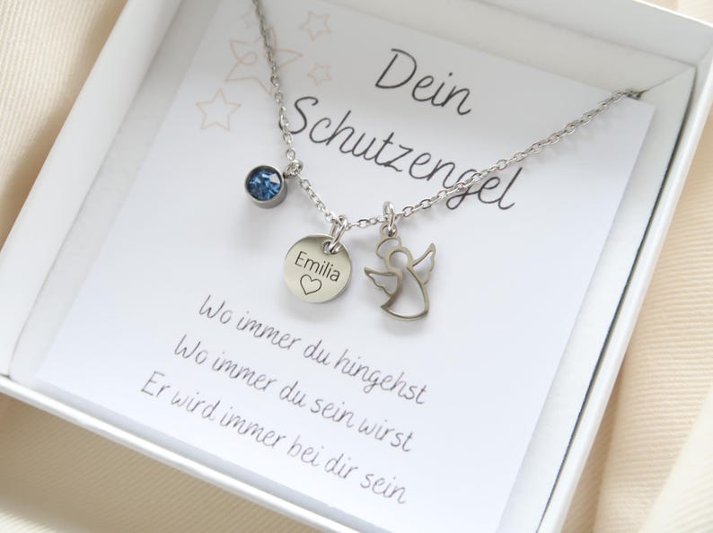 May include: A silver necklace with three charms, presented in a white box. The charms include a blue gemstone, a round pendant with the name "Emilia" and a heart, and a small angel. The card reads "Dein Schutzengel".