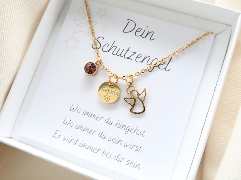 May include: A gold-colored necklace with three charms, including a round pendant engraved with "Sophia" and a heart, an angel outline, and a purple gemstone. The necklace is presented in a white box with German text.
