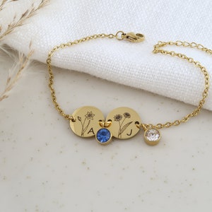 May include: Gold-toned bracelet featuring two engraved circular charms with floral designs and the letters "A" and "J". The bracelet includes a blue and a clear crystal charm. The chain is delicate and has a lobster clasp.