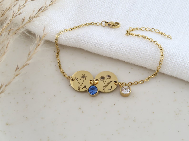 May include: Gold-toned bracelet featuring two engraved circular charms with floral designs and the letters "A" and "J". The bracelet includes a blue and a clear crystal charm. The chain is delicate and has a lobster clasp.