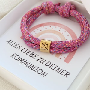 Könnte beinhalten: Ein rosa-mehrfarbiges Seilarmband mit einer goldfarbenen zylindrischen Perle, die mit dem Namen "Ida" und einem Fischsymbol graviert ist. Das Armband wird in einer weißen Schachtel mit einer Karte mit einer Regenbogenillustration und deutschem Text präsentiert.