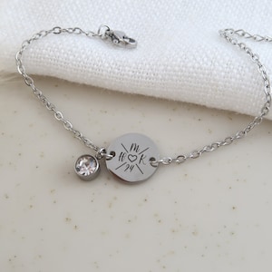 May include: A silver-colored bracelet with a delicate chain. It features a round charm with engraved initials and a date, along with a small, clear crystal. The bracelet is displayed on a white surface.