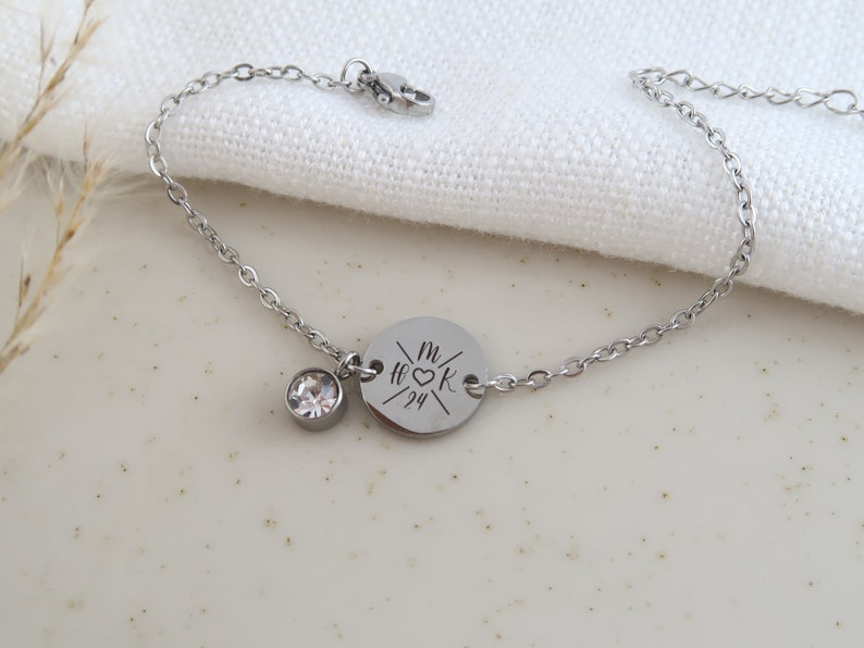 May include: A silver-colored bracelet with a delicate chain. It features a round charm with engraved initials and a date, along with a small, clear crystal. The bracelet is displayed on a white surface.