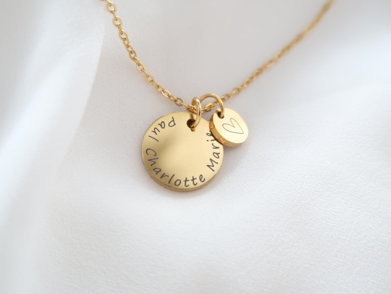 May include: A gold-colored necklace with two circular pendants. The larger pendant has the names "Paul" and "Charlotte" engraved around the edge. The smaller pendant has the name "Marie" and a heart symbol. The necklace is on a white background.