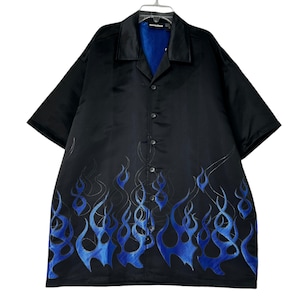 Puede incluir: Camisa negra de manga corta con cuello de solapa y diseño de llamas azules. La camisa tiene forro azul y está colgada de una percha blanca. La camisa está abotonada.