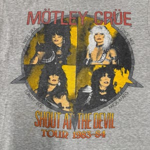 以下が含まれることがあります： Mötley CrüeのグラフィックがプリントされたグレーのTシャツ。バンド名、メンバー名、そして「SHOUT AT THE DEVIL TOUR 1983-84」の文字が赤と黄色でデザインされています。