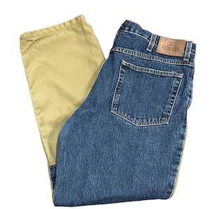 Puede incluir: Vaqueros de mezclilla azules con un panel de color beige en el lateral. Los vaqueros tienen un diseño clásico de cinco bolsillos y un parche de cuero con el nombre de la marca "RedHead" en la parte trasera. Los vaqueros están doblados.