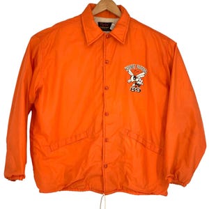 May include: Bright orange vintage jacket with a button-up front, collar, and drawstring hem. The jacket features a Hornet Football logo with a cartoon hornet and the number 89 on the chest. The jacket is hanging on a wooden hanger.