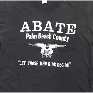 May include: Black t-shirt with white text and a pocket. The shirt features the word "ABATE" above "Palm Beach County" and an emblem of an eagle with a shield. The phrase "LET THOSE WHO RIDE DECIDE" is printed below.
