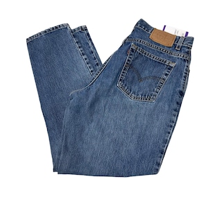May include: Blue denim jeans with a classic five-pocket design. The jeans feature a brown leather patch on the back and a small tag. The jeans are folded and displayed on a white background.