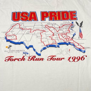 May include: White t-shirt featuring a map of the USA with a red route, the words "USA PRIDE," and "Torch Run Tour 1996." The shirt also includes the text "All Over America" and a stylized eagle.