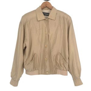 Puede incluir: Chaqueta de color beige con cuello y cierre de botones. La chaqueta tiene dos bolsillos delanteros y un dobladillo y puños acanalados. La etiqueta dice "LEE GILMAN". La chaqueta está colgada de una percha de madera.