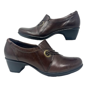 Op de afbeelding: Twee bruine leren schoenen met een lage hak. Elke schoen heeft een decoratief metalen ringdetail op de bovenkant en een zwarte zool. De schoenen worden tegen een witte achtergrond getoond.