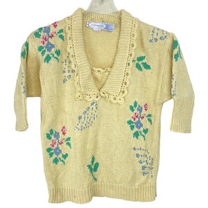 Könnte beinhalten: Ein hellgelber Pullover mit V-Ausschnitt und kurzen Ärmeln. Der Pullover hat ein Blumenmuster in Rosa, Blau und Grün. Der Kragen und der Ausschnitt sind mit einem zarten Spitzendesign verziert. Das Etikett lautet "J. Christopher".