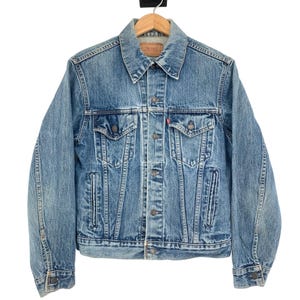May include: A light blue denim jacket with a classic design. The jacket features a button-up front, two chest pockets, and two side pockets. The jacket is hanging on a wooden hanger.