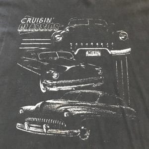 May include: Dark gray t-shirt featuring a graphic of classic cars in white. The text "Cruisin' Classics" is above the car illustrations. The design includes three vintage cars, with the word "MERC" on a license plate.