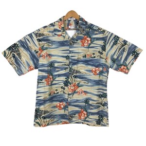 Puede incluir: Camisa de manga corta con botones y estampado de paisaje tropical. La camisa presenta un fondo ondulado azul y beige con flores naranjas y palmeras. La camisa está colgada de una percha de madera.