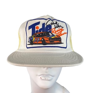 May include: Cream-coloured Tide Racing Team trucker hat. The front panel features a Tide logo with a race car and the number 17. The hat has a mesh back and a curved brim. The hat also has a signature on the front.