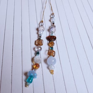 Könnte beinhalten: Zwei Perlenornamente mit einer Vielzahl farbenfroher Perlen. Die Perlen umfassen klare, braune, weiße und blaue Perlen, von denen einige wie Schmetterlinge und andere einzigartige Designs geformt sind. Die Ornamente hängen an dünnen Goldfäden.